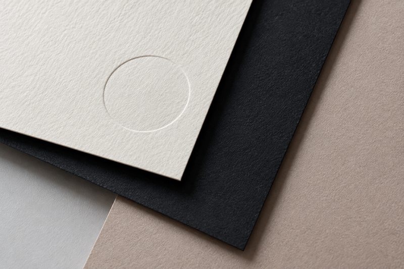 Layered sheets of warm white, black and beige uncoated card; the top sheet carries a debossed ring.